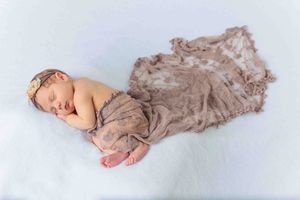 Peaceful newborn baby sleeping wrapped in a soft taupe lace swaddle with floral headband.