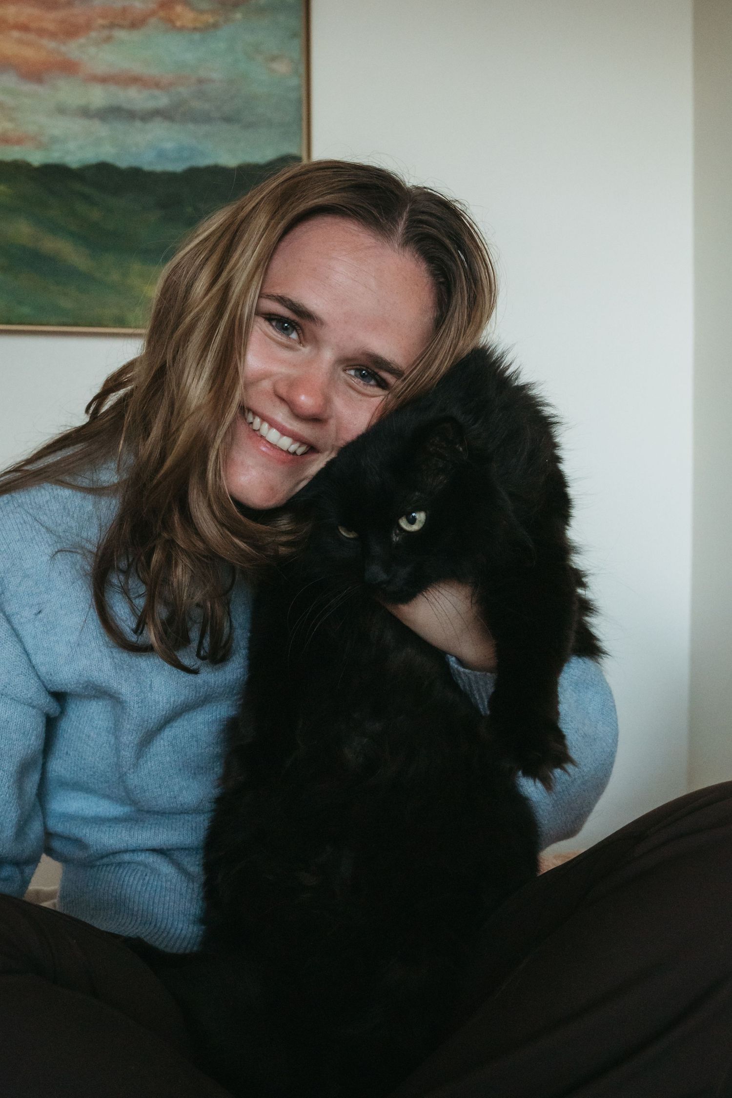 Smiling woman in blue sweater holding a fluffy black cat close to her face indoors.