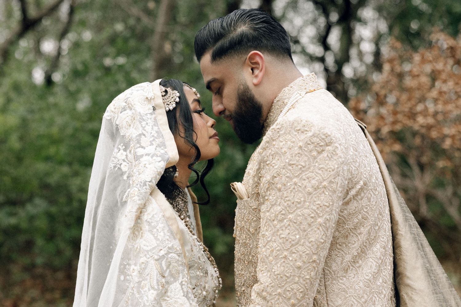 A couple in intricate cream-colored traditional wedding attire share an intimate moment in a garden setting.