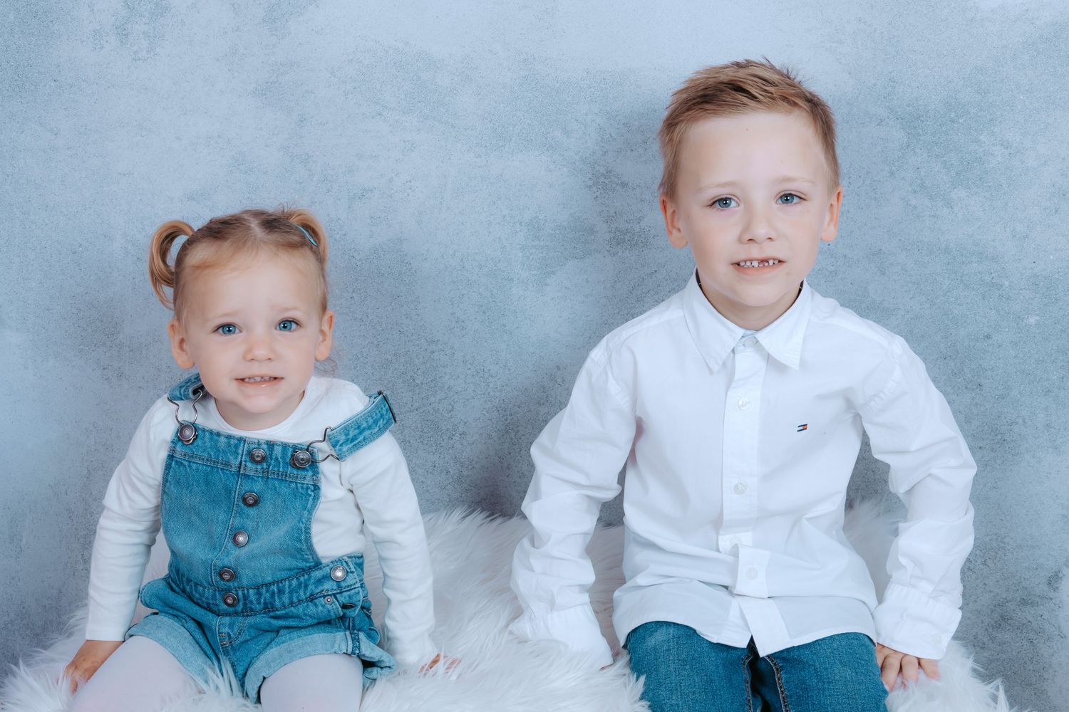Two young children, a toddler girl in denim overalls and a boy in a white shirt, sitting together for a portrait.