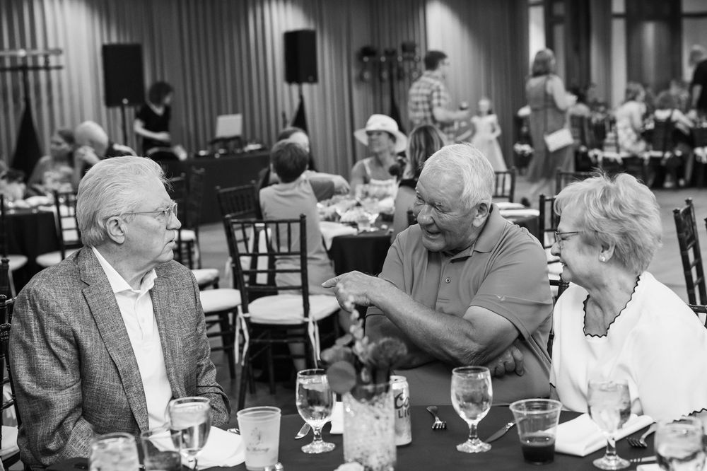 Three elderly people engaged in conversation at a formal banquet table with wine glasses in a decorated event hall.