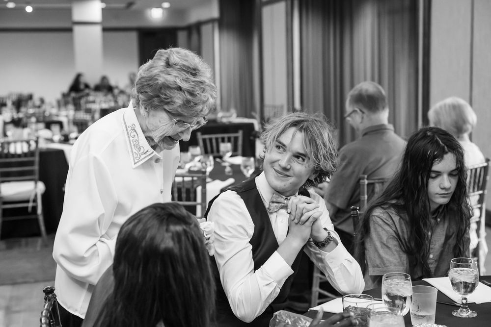 Black and white photo of guests mingling at a formal banquet event, with a young man in a vest smiling warmly.