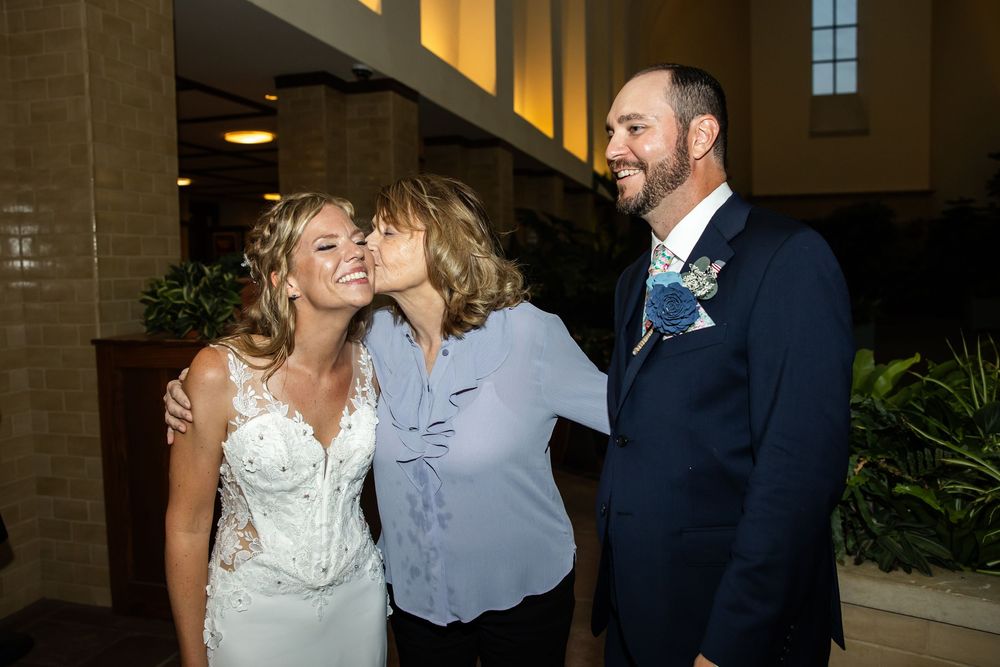 Bride in white lace gown receives a kiss on the cheek while groom in navy suit smiles at a wedding reception.