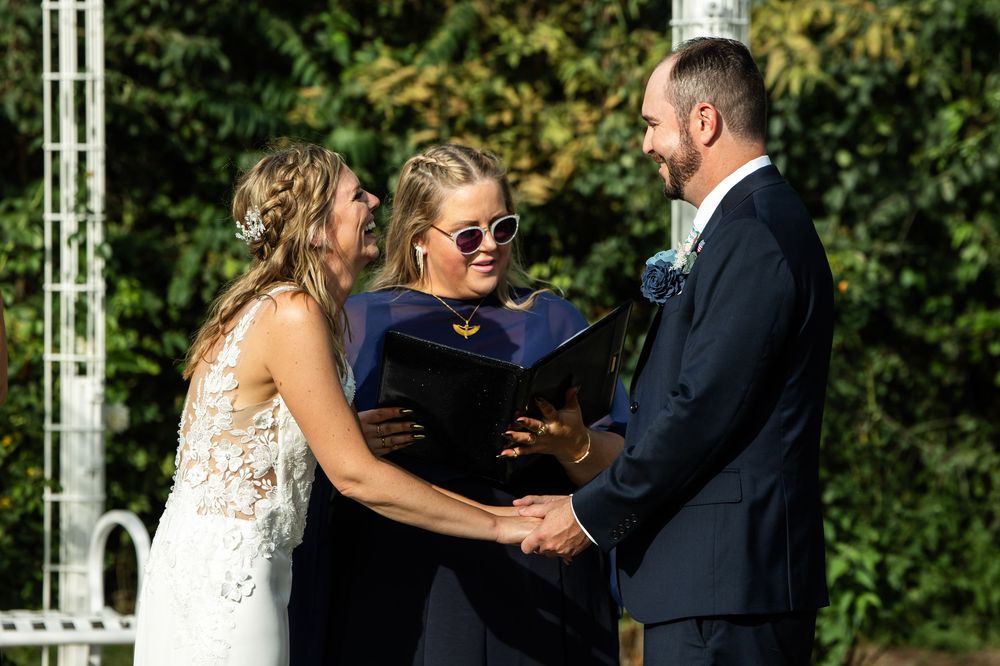 Outdoor wedding ceremony with bride and groom holding hands before officiant in navy graduation gown and sunglasses.