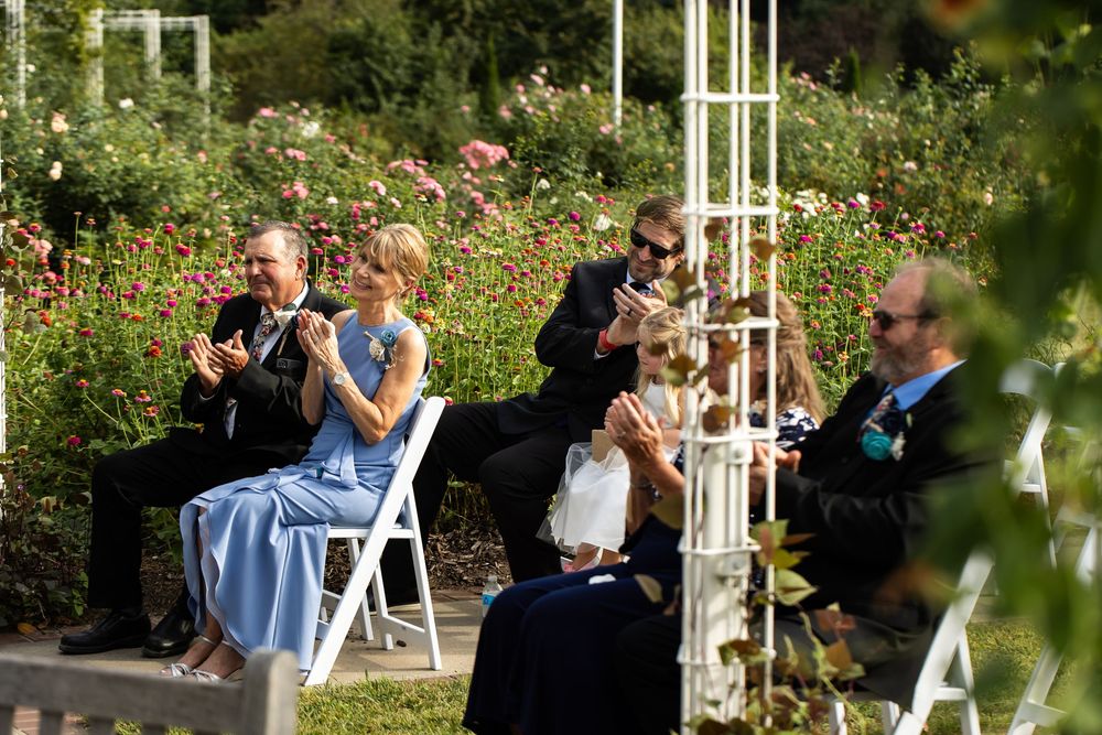 Guests clapping and seated on white chairs at an outdoor wedding ceremony surrounded by blooming rose gardens.