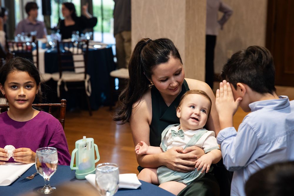 A woman holds a laughing baby at a restaurant table while a boy waves and a girl smiles nearby.
