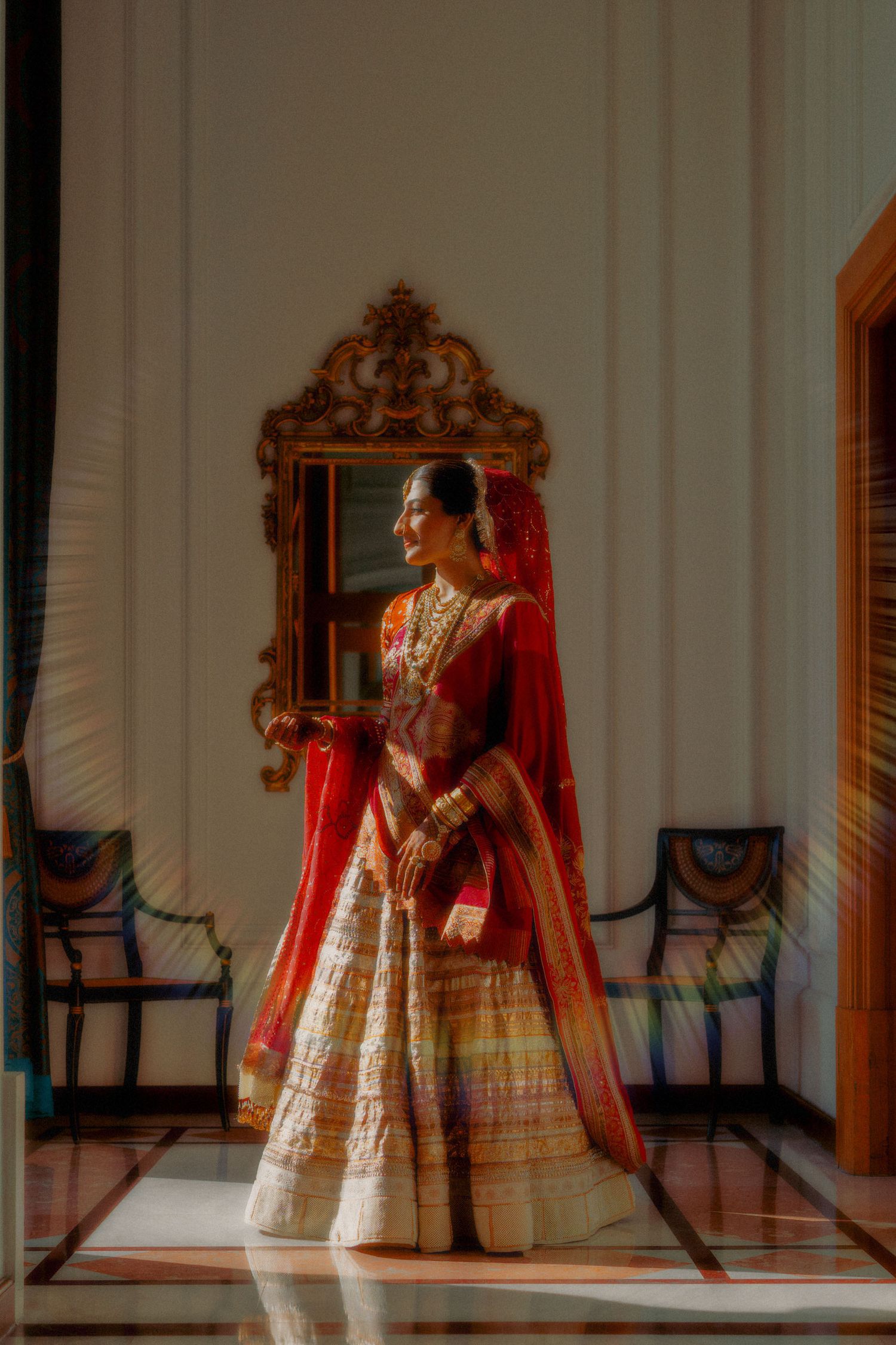 Indian bride in red and gold lehenga standing in sunlit room with ornate mirror, traditional bridal jewelry and dupatta.