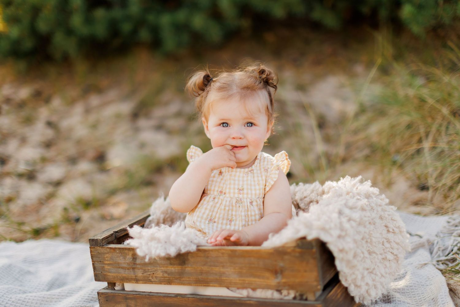 Smiling baby girl in yellow romper sitting in rustic wooden crate outdoors with fluffy blanket.