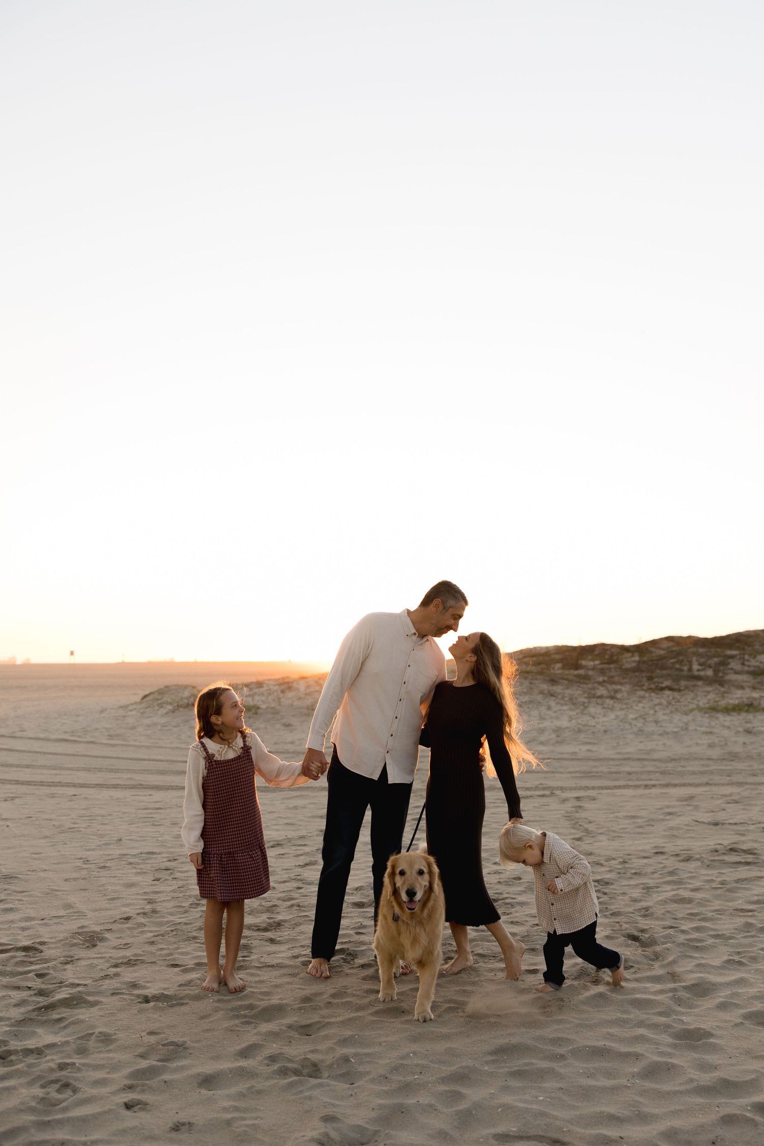 Family with two children and golden retriever dog posing on sandy beach at sunset.