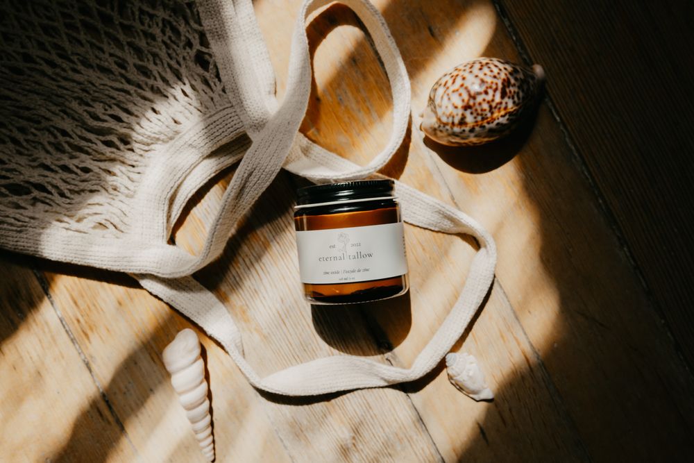 Amber glass candle jar beside a decorative shell and mesh bag on a sunlit wooden surface.