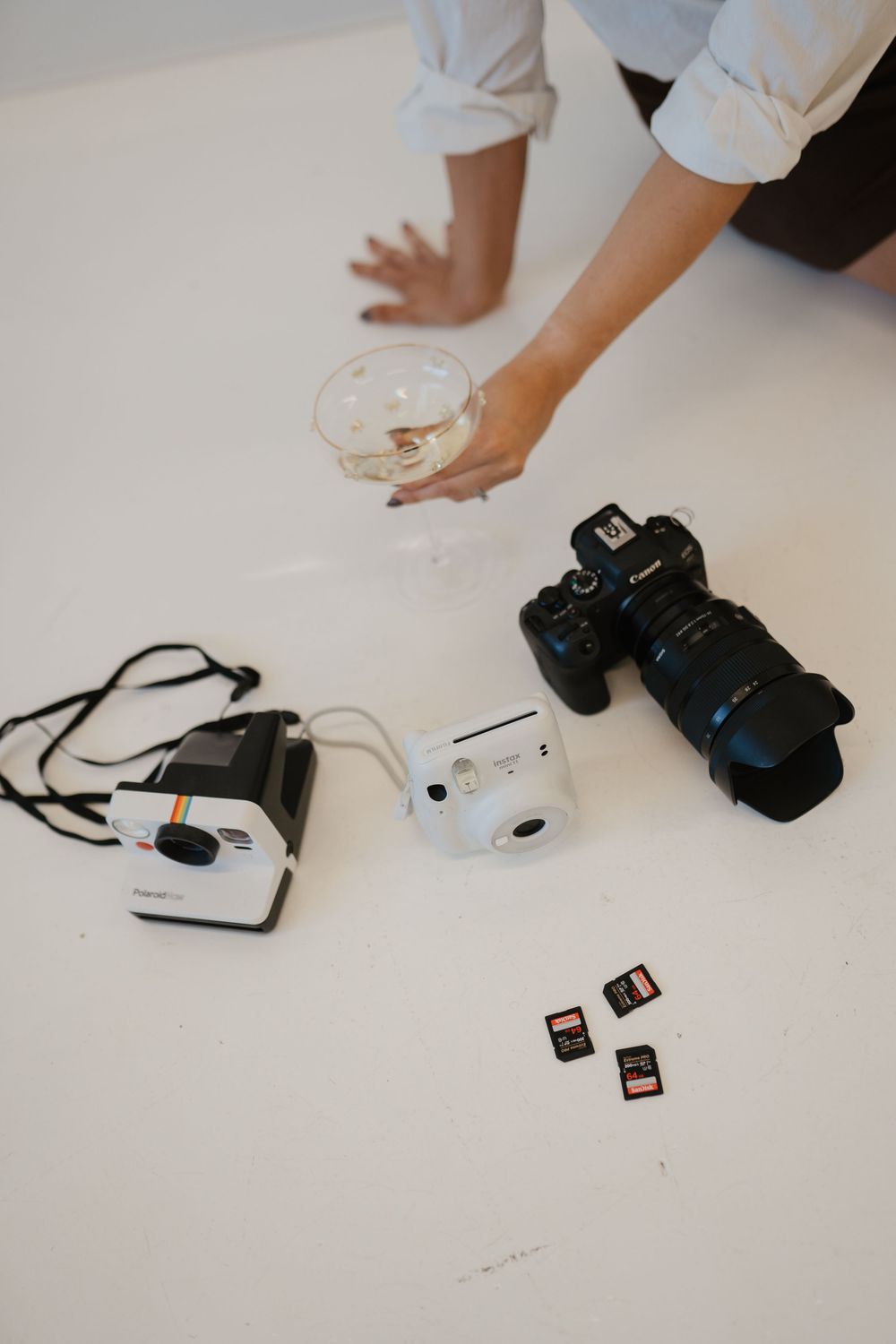 Person reaching for a wine glass beside a Polaroid, Instax Mini, Canon DSLR camera, and SD cards on white surface.