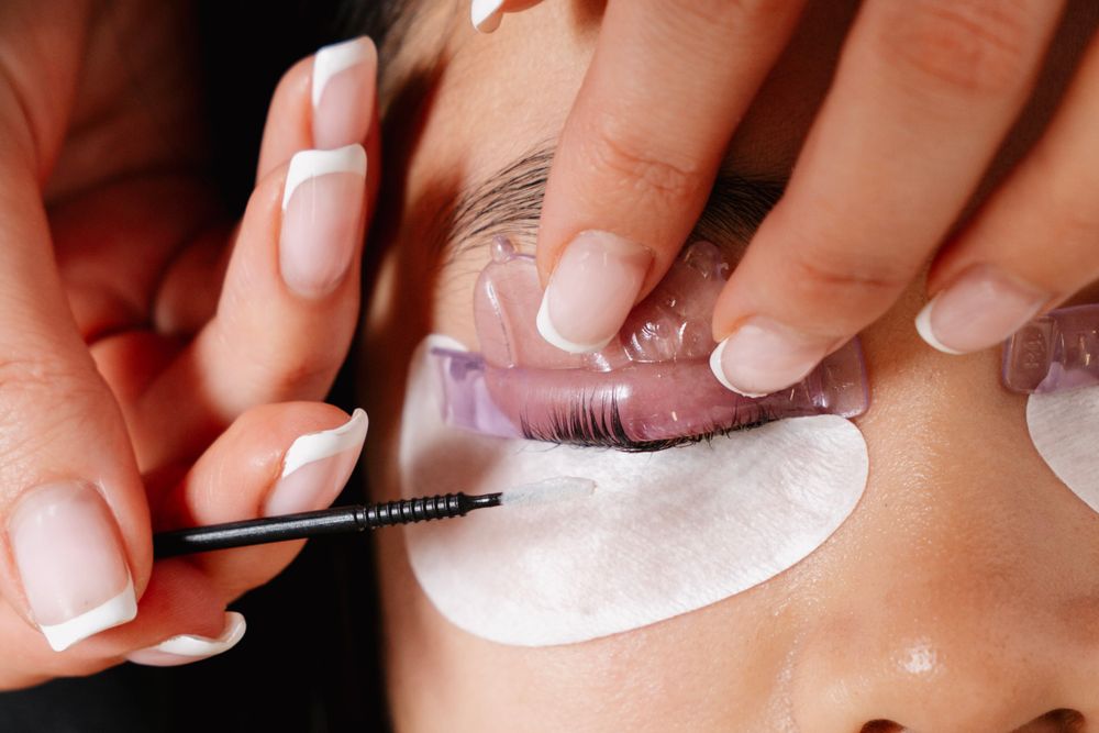 Close-up of eyelash extension application with a lash brush on a client with white under-eye pads.