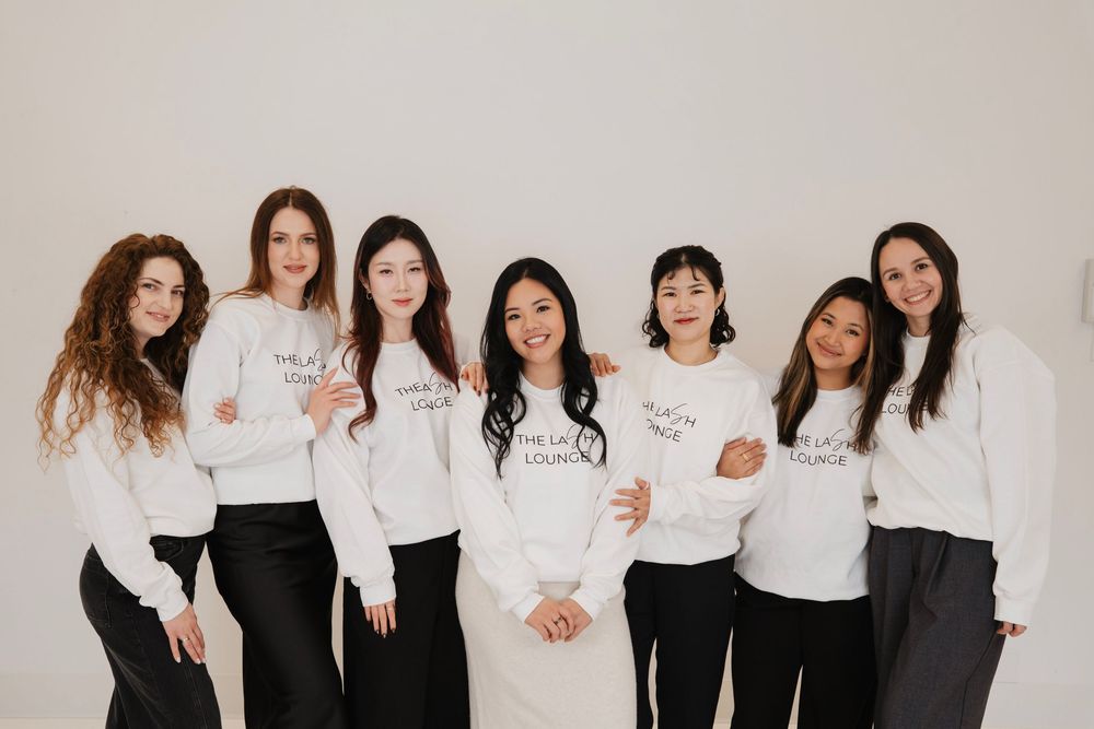 Seven women in matching white sweatshirts and black pants pose together smiling against a white background.