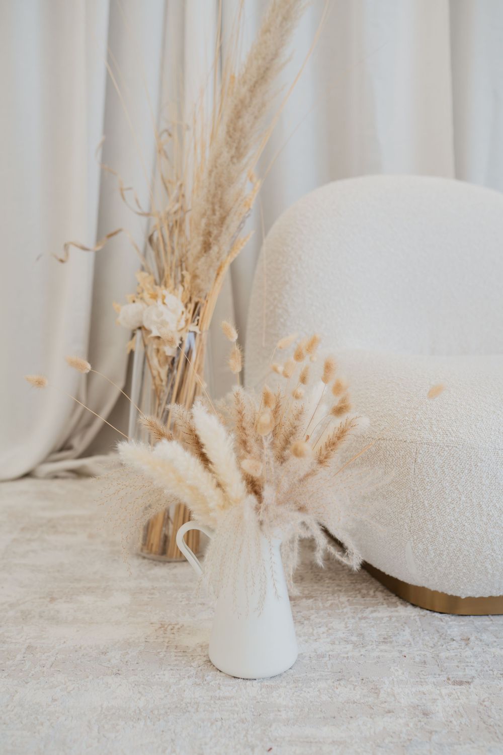 White vase with dried pampas grass beside a fluffy cream chair and curtains in a minimalist boho interior.