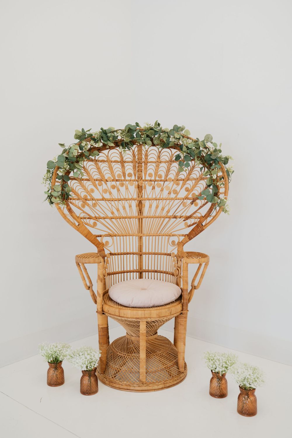 Elegant peacock rattan chair with greenery garland and baby's breath in copper vases on white background.