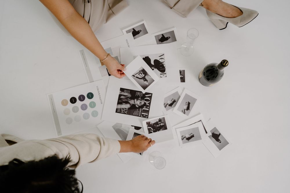Two people reviewing black and white photos spread on a white table with color swatches and a glass bottle.