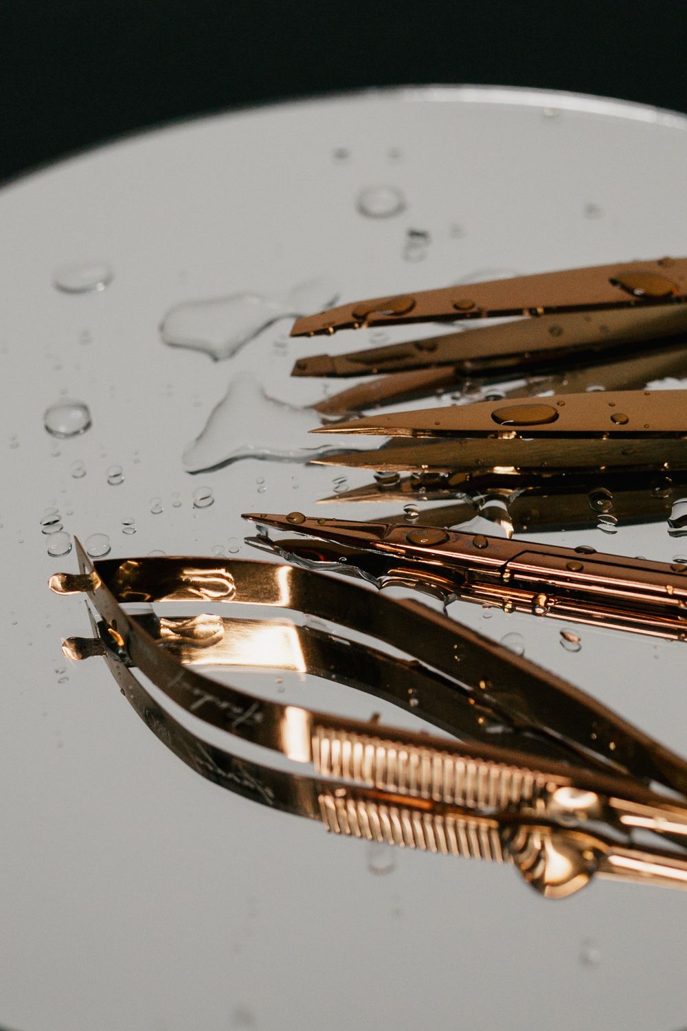 Gold rose-toned tweezers and nail scissors arranged on a wet reflective white surface with water droplets.
