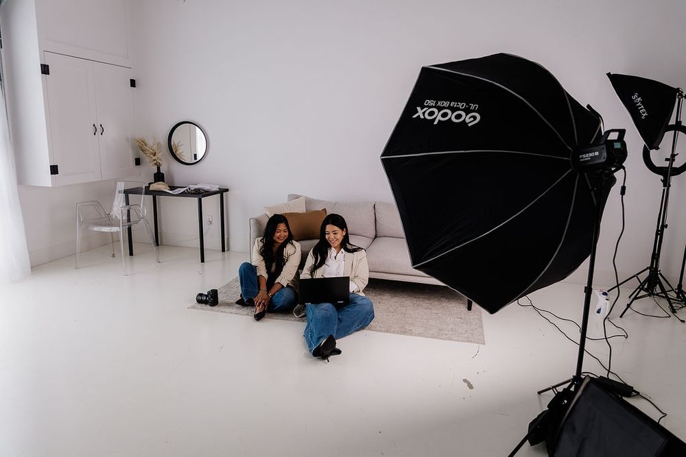 Two women sit on the floor of a professional photo studio with Godox umbrella lighting equipment and white walls.
