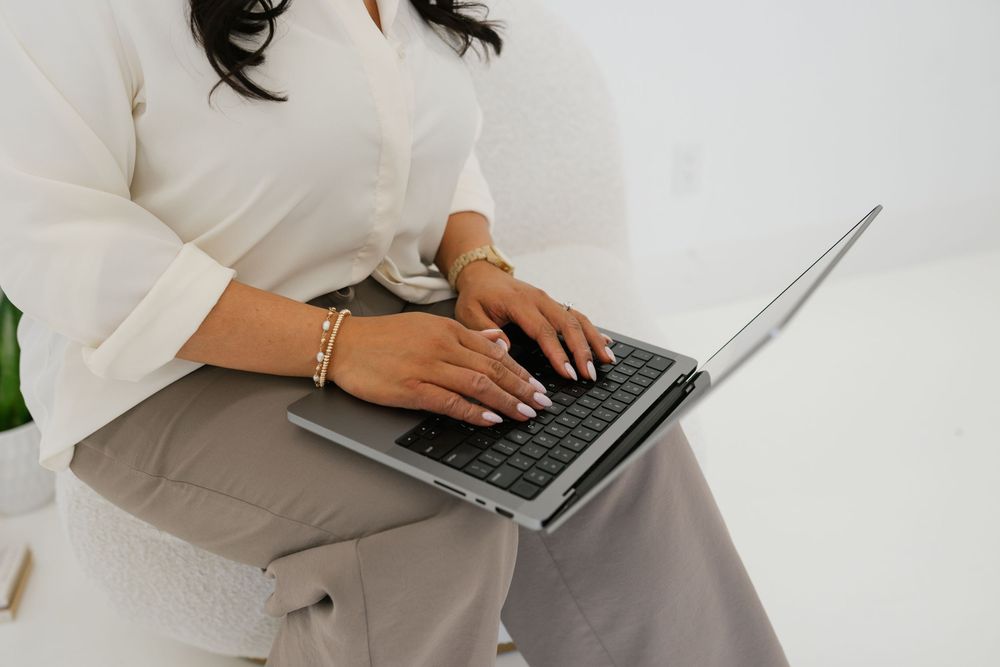 Woman in white blouse and gray pants typing on a silver laptop while seated, wearing gold bracelets.
