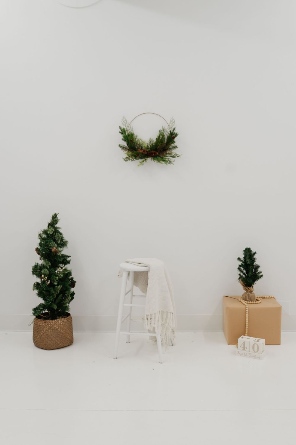 Minimalist Christmas decor with two small pine trees, a white stool, wreath, and wrapped gift on white background.