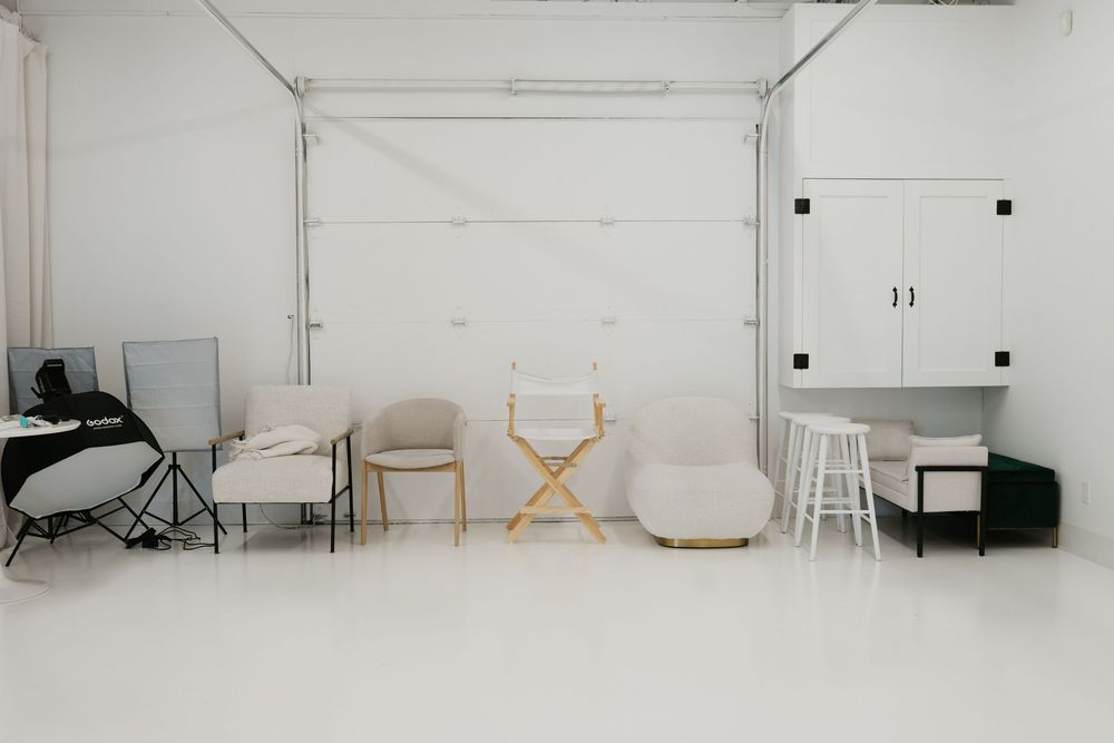 Bright white photography studio with assorted chairs, director's chair, bar stools, and white cabinets in minimalist setup.