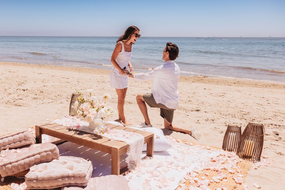 A romantic beach proposal scene with decorative pillows and woven baskets on the sand.