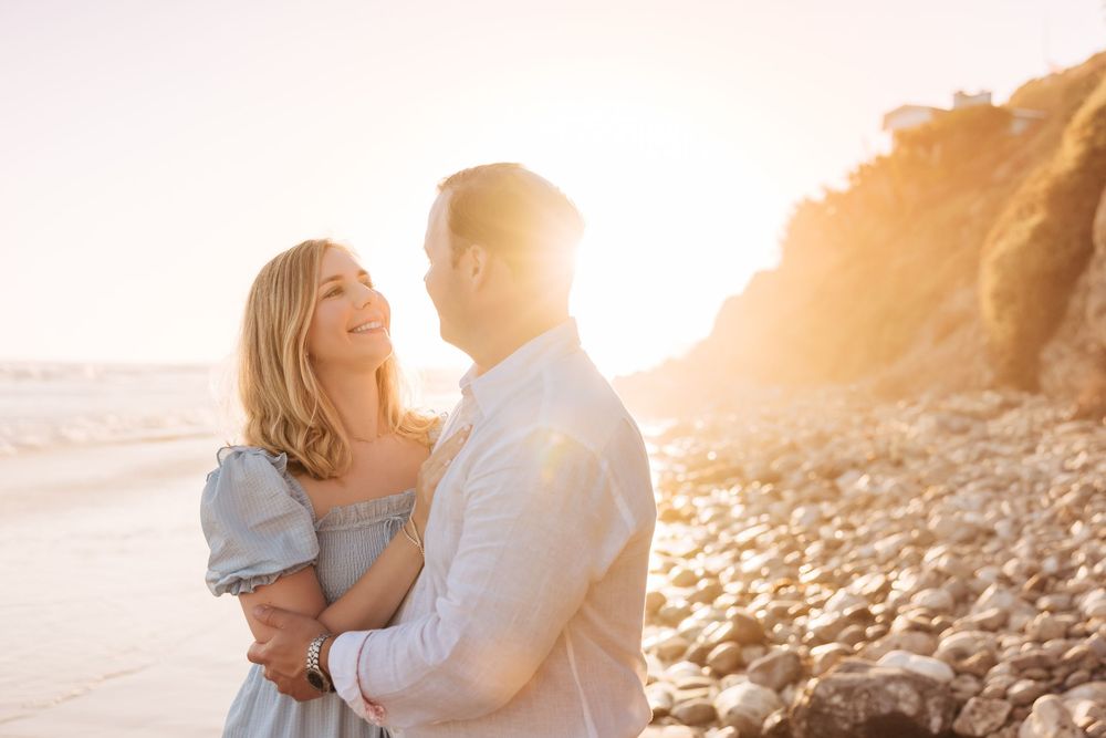 Couple embracing on a rocky beach at golden sunset, woman in blue dress smiling up at man in white shirt.