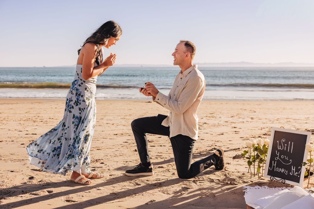 Man proposing on one knee to woman on a sunny beach, holding a ring with a will you marry me sign nearby.