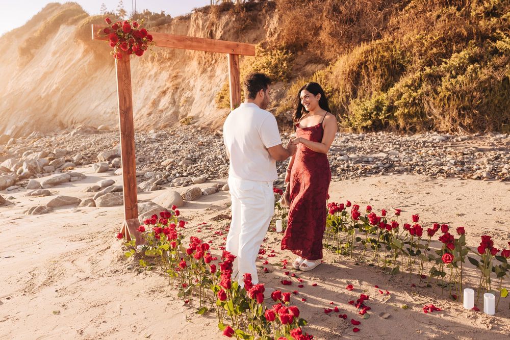 Couple sharing a romantic beach proposal surrounded by red roses under a wooden arch at golden hour.