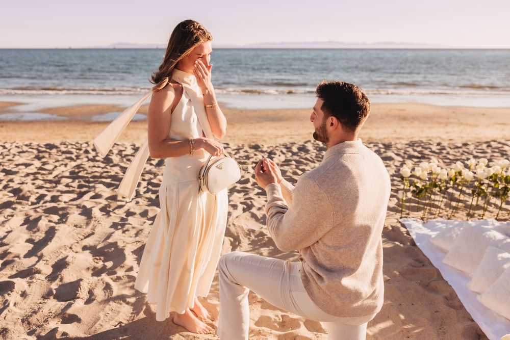 Man proposes to woman on a sandy beach at sunset, holding a ring while she covers her face in surprise.