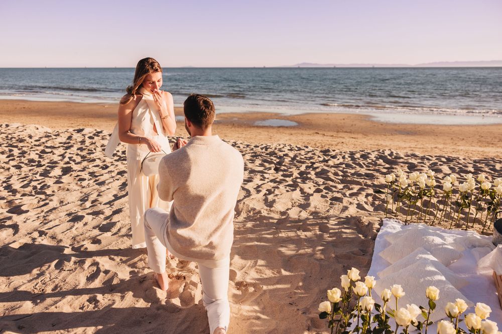 Man kneeling on sandy beach proposing to woman near ocean with white roses and romantic sunset backdrop.