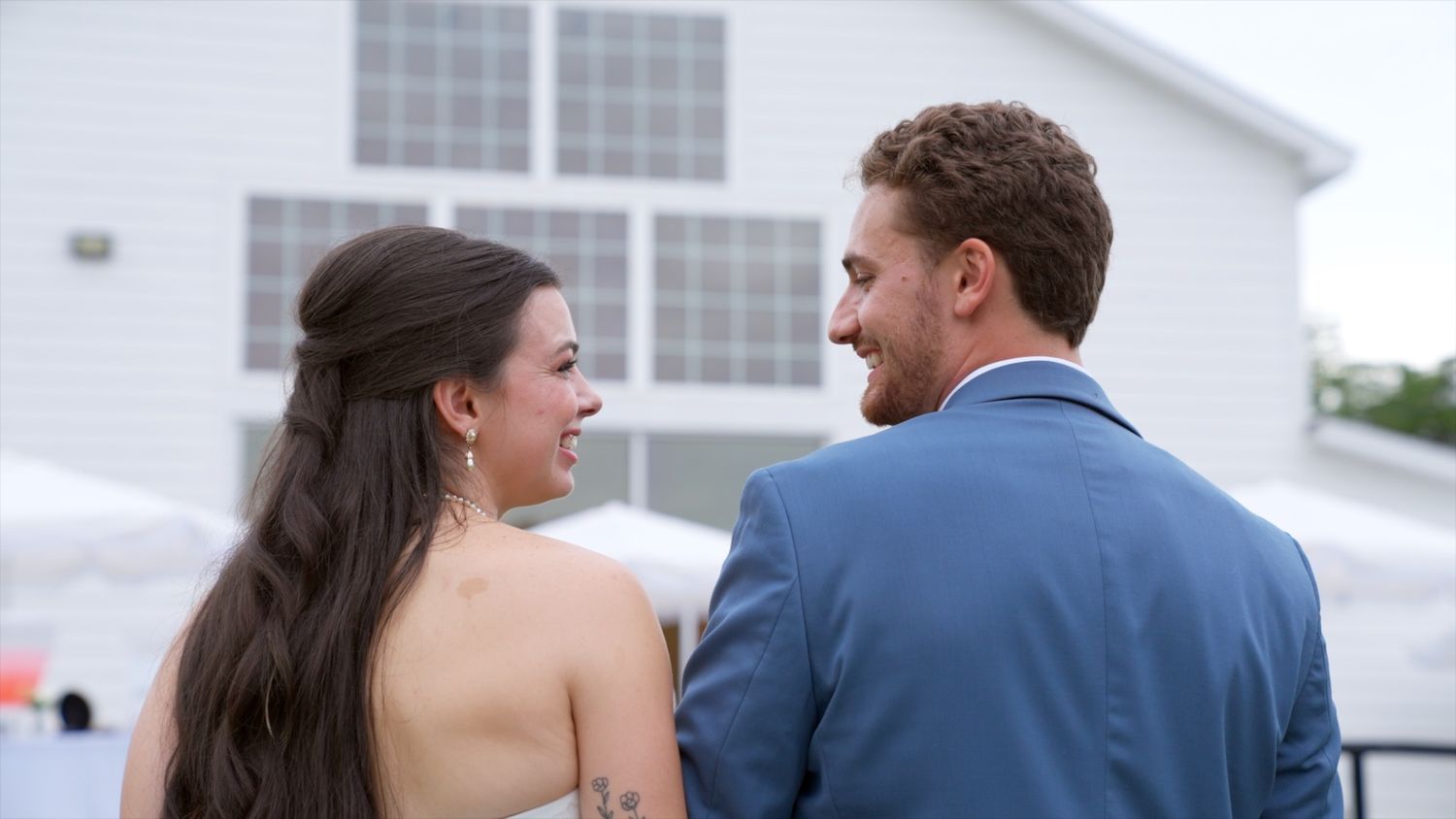 A couple in wedding attire smiles at each other outdoors near a white building on their special day.