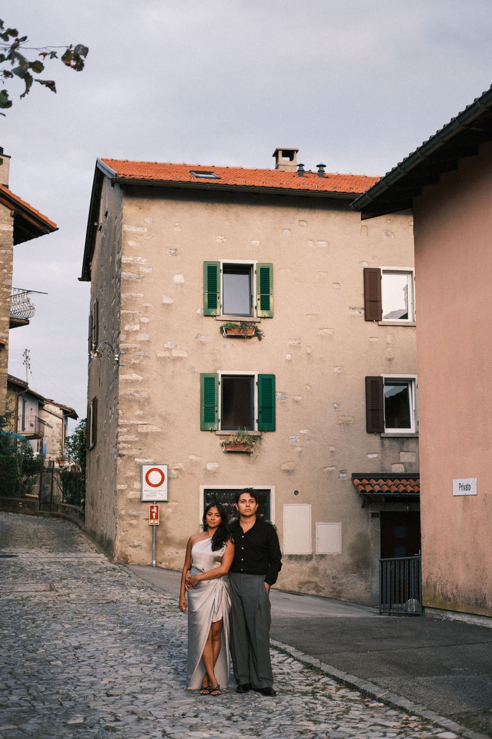 Couple embracing on cobblestone street in front of rustic European building with green shutters.