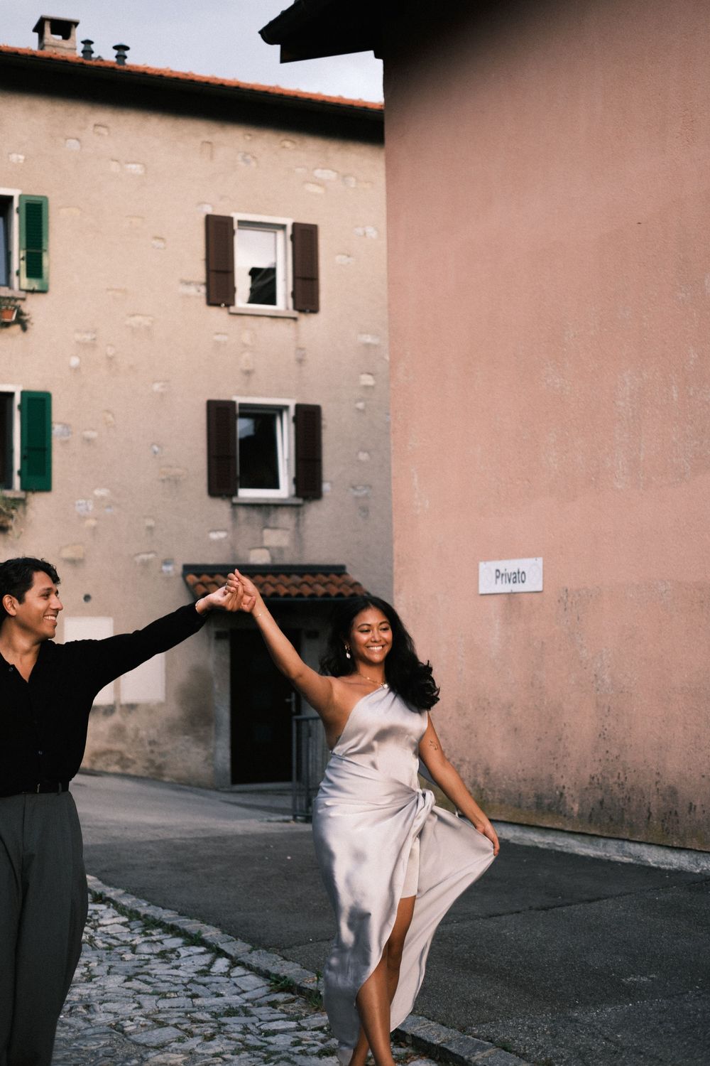 Couple dancing joyfully on a cobblestone street in an Italian village, woman in silver dress, man in black shirt.