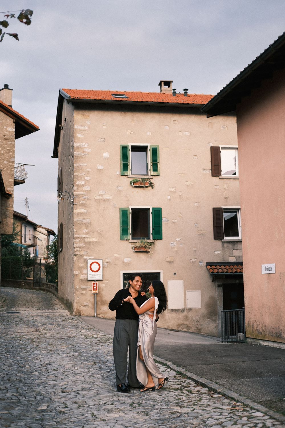 Couple dancing on cobblestone street in charming European village with colorful shuttered buildings backdrop.