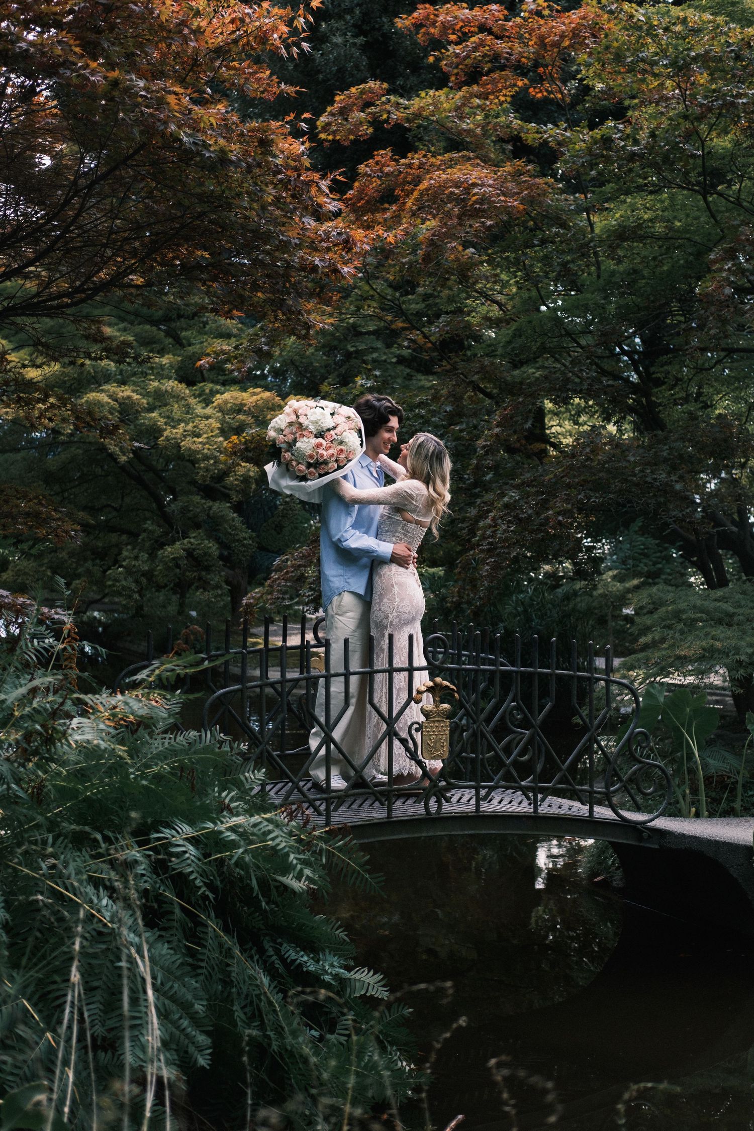 Couple embracing on ornate bridge surrounded by autumn foliage in a lush garden, holding a large floral bouquet.