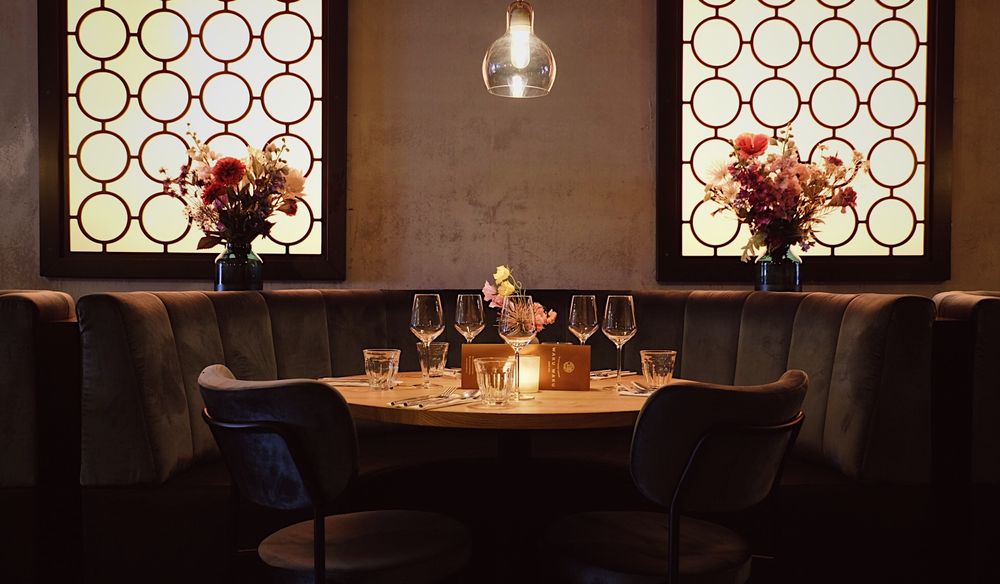 Elegant restaurant booth with velvet seating, round table, wine glasses, candlelight, and ornate backlit windows with floral arrangements.