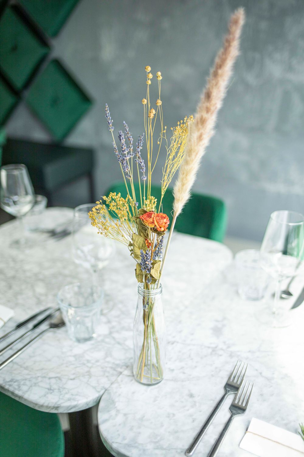 Elegant marble table with dried flower arrangement in glass vase, green velvet chairs, modern restaurant setting.