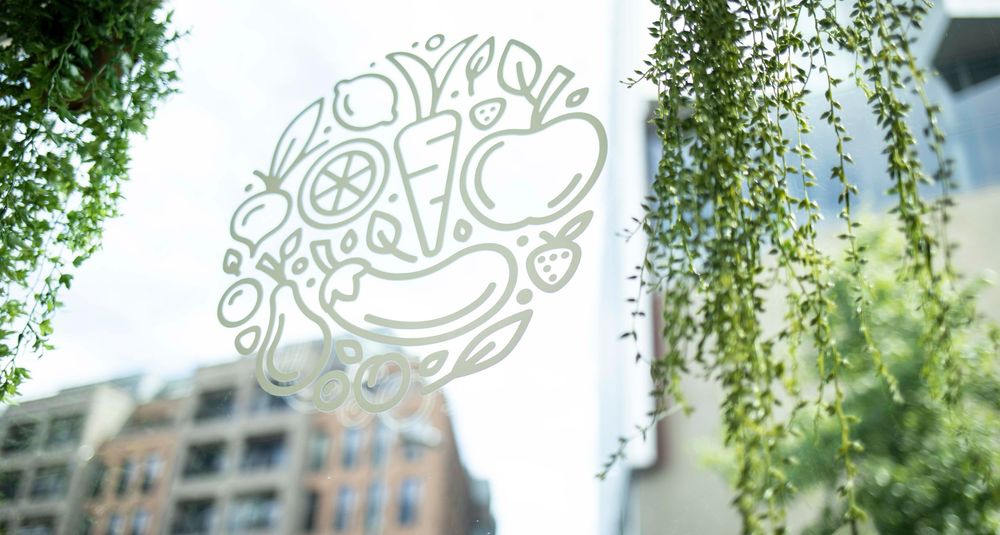 White illustrated food logo on glass window surrounded by lush green plants with urban buildings in background.