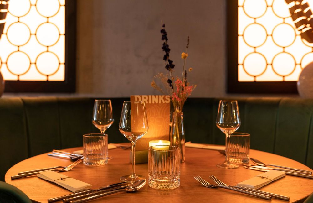 Elegant restaurant table set for two with wine glasses, candle, and drinks menu centerpiece in warm ambient lighting.