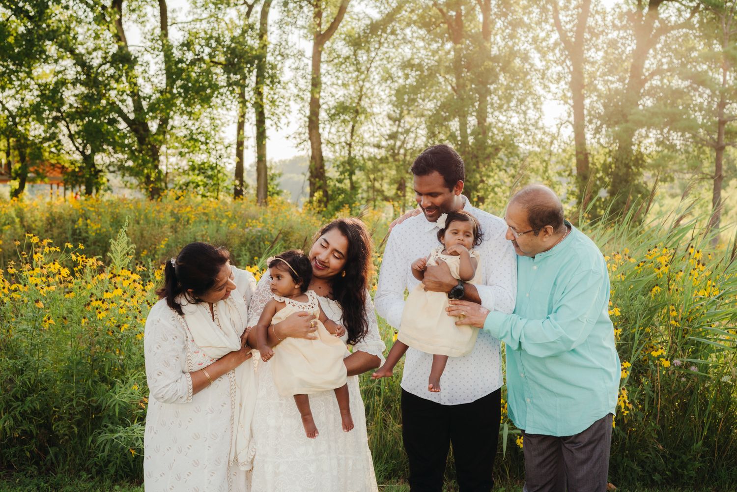 Multigenerational family in white and soft colors holds two babies and shares a warm moment in a sunlit field of yellow wildflowers.
