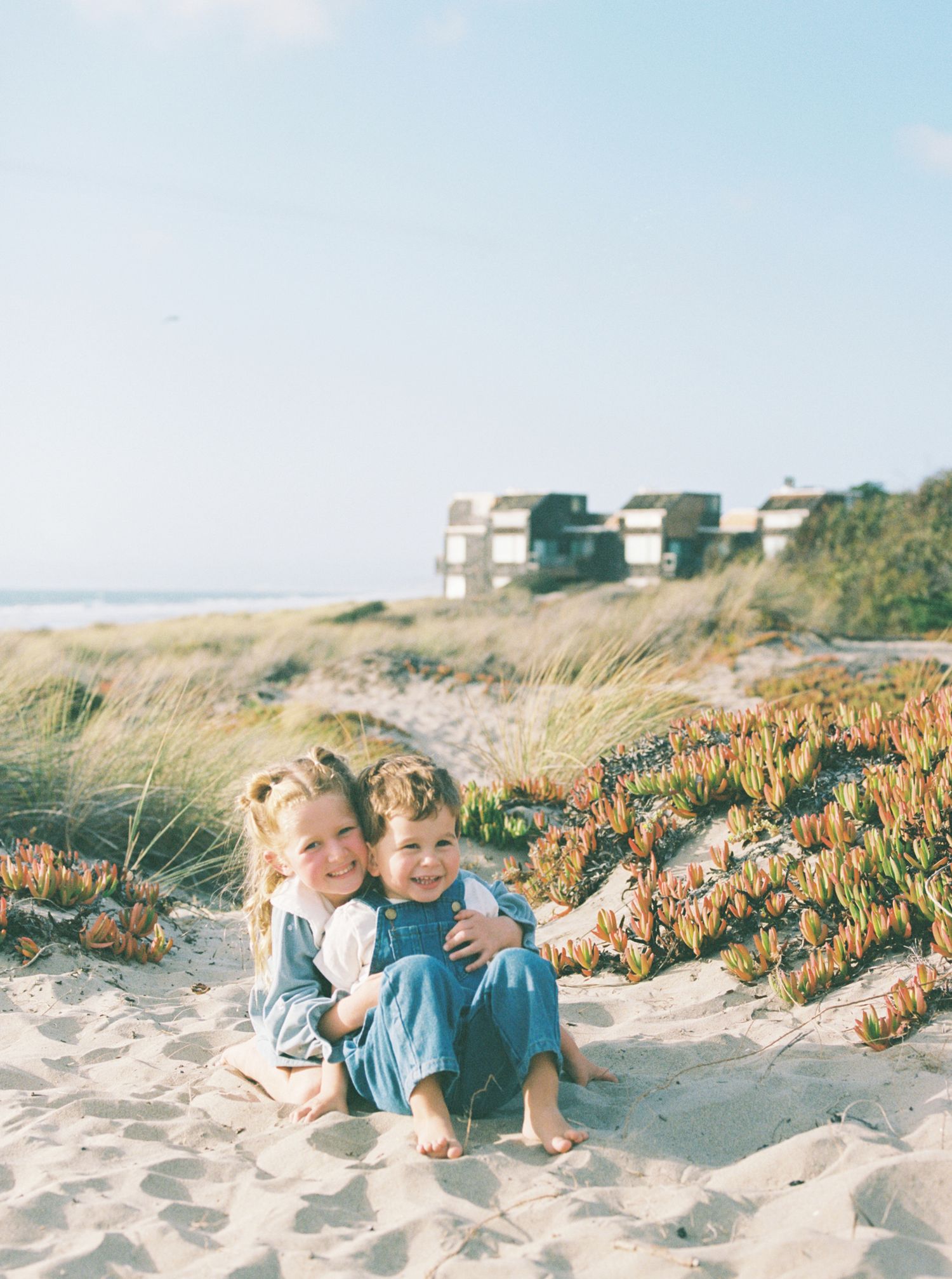 Two smiling young children hug while sitting barefoot on a sandy beach dune with ice plant and dune grass near coastal houses.