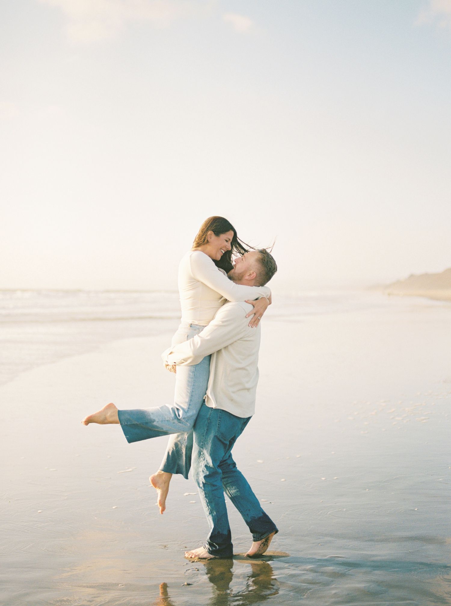 Barefoot couple embracing on a sunlit beach as the man lifts the smiling woman off the ground at the water's edge.