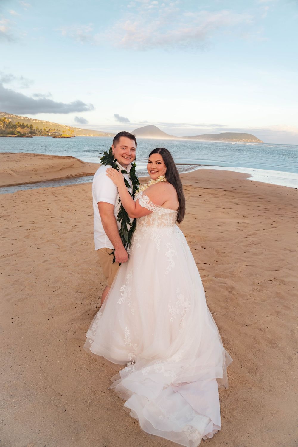 Smiling bride in a white lace wedding gown and groom wearing a green leaf lei embrace on a sandy beach with ocean and hills behind them.