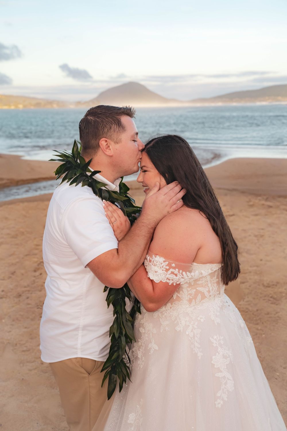 Groom in white shirt and green maile lei kisses bride in off-shoulder lace wedding gown on forehead during a beach wedding at sunset.