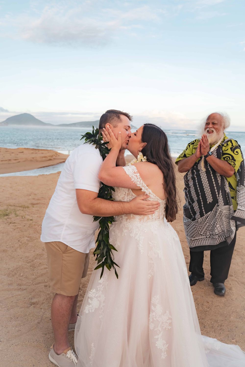 Bride in a lace wedding gown kisses her groom wearing a green leaf lei as an officiant applauds during a beach wedding in Hawaii.