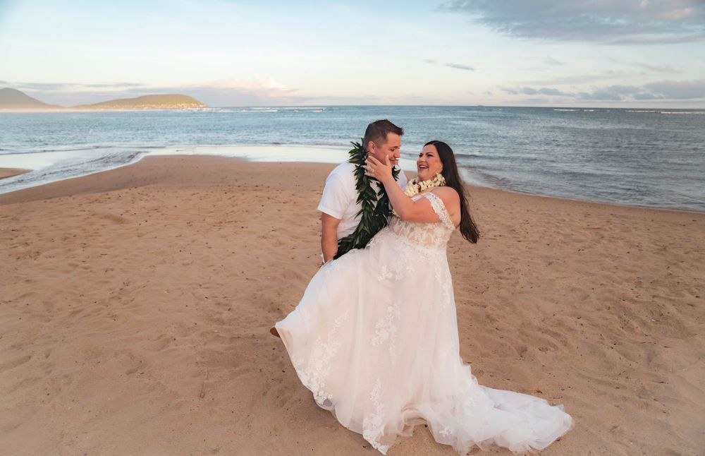 Bride in a flowing white gown and groom wearing a green leaf lei embrace on a sandy beach at sunset with the ocean behind them.