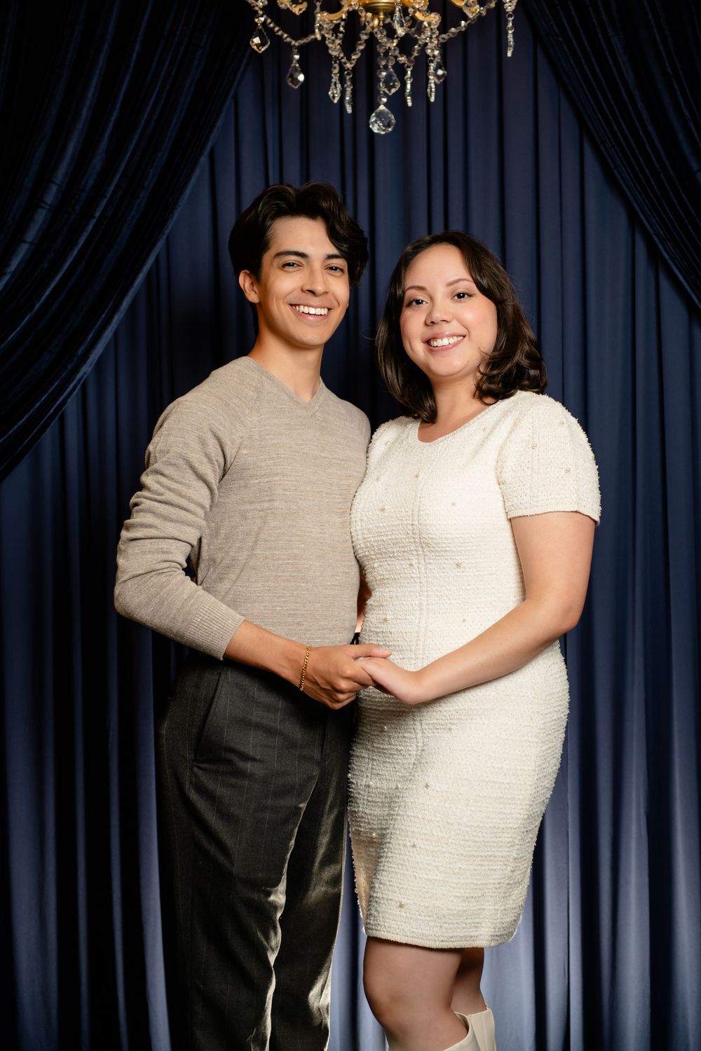 Smiling couple holding hands in a cozy pose in front of deep blue velvet curtains under a crystal chandelier.