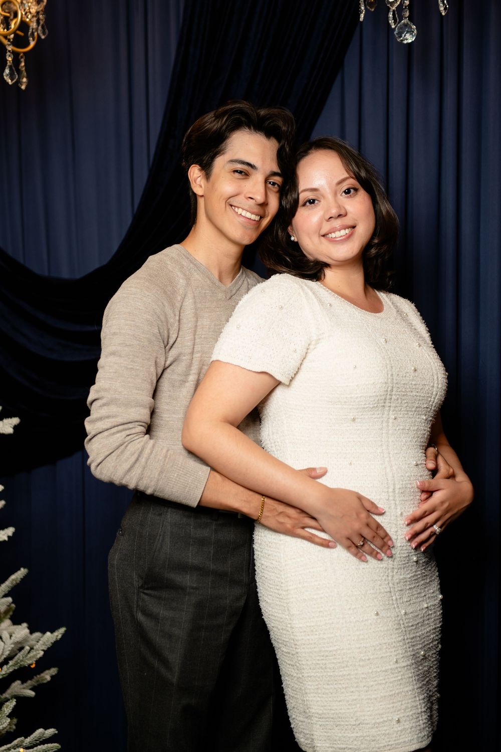 Smiling couple in a maternity pose, the woman in a white dress with hands on her baby bump, against a navy curtain backdrop.