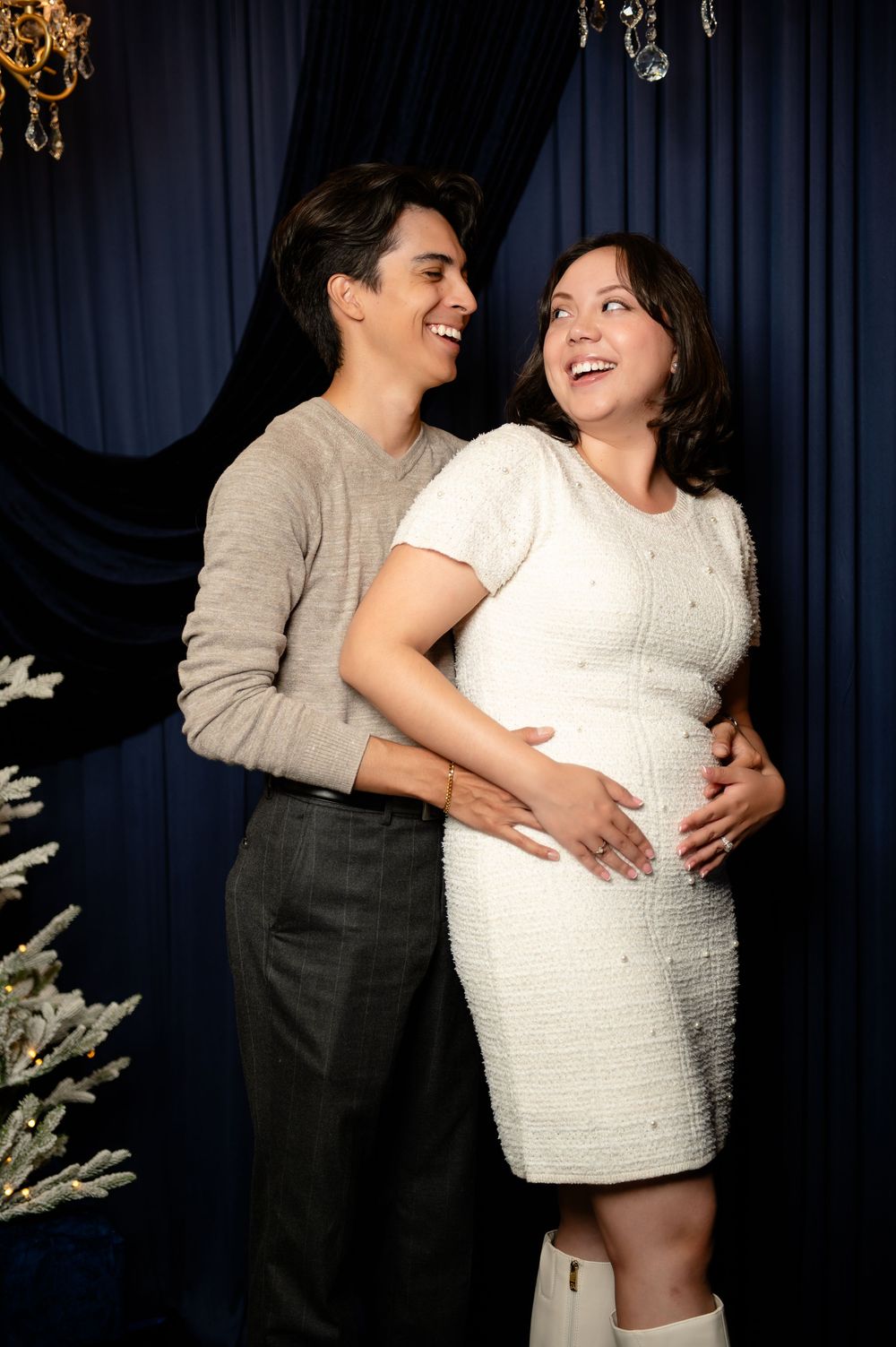 Smiling man embraces a laughing pregnant woman in a white dress, her hands on her baby bump, against a navy curtain.